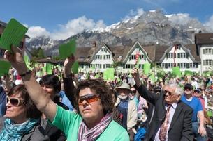 Voters hold up green cards with traditional buildings and houses in background