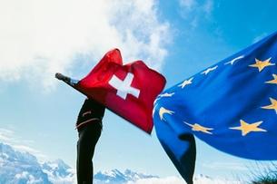 Schweizer- und EU-Flagge wehen vor blauem Himmel. Im Hintergrund schneebedeckte Berge 